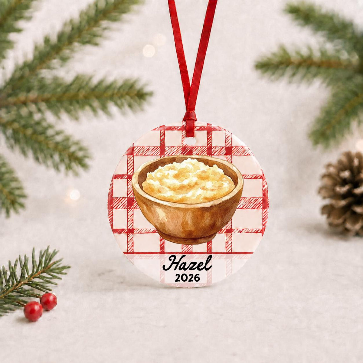 Personalized mashed potatoes ceramic ornament with name and year on red plaid with red ribbon on a white surface with pine branches and pinecones