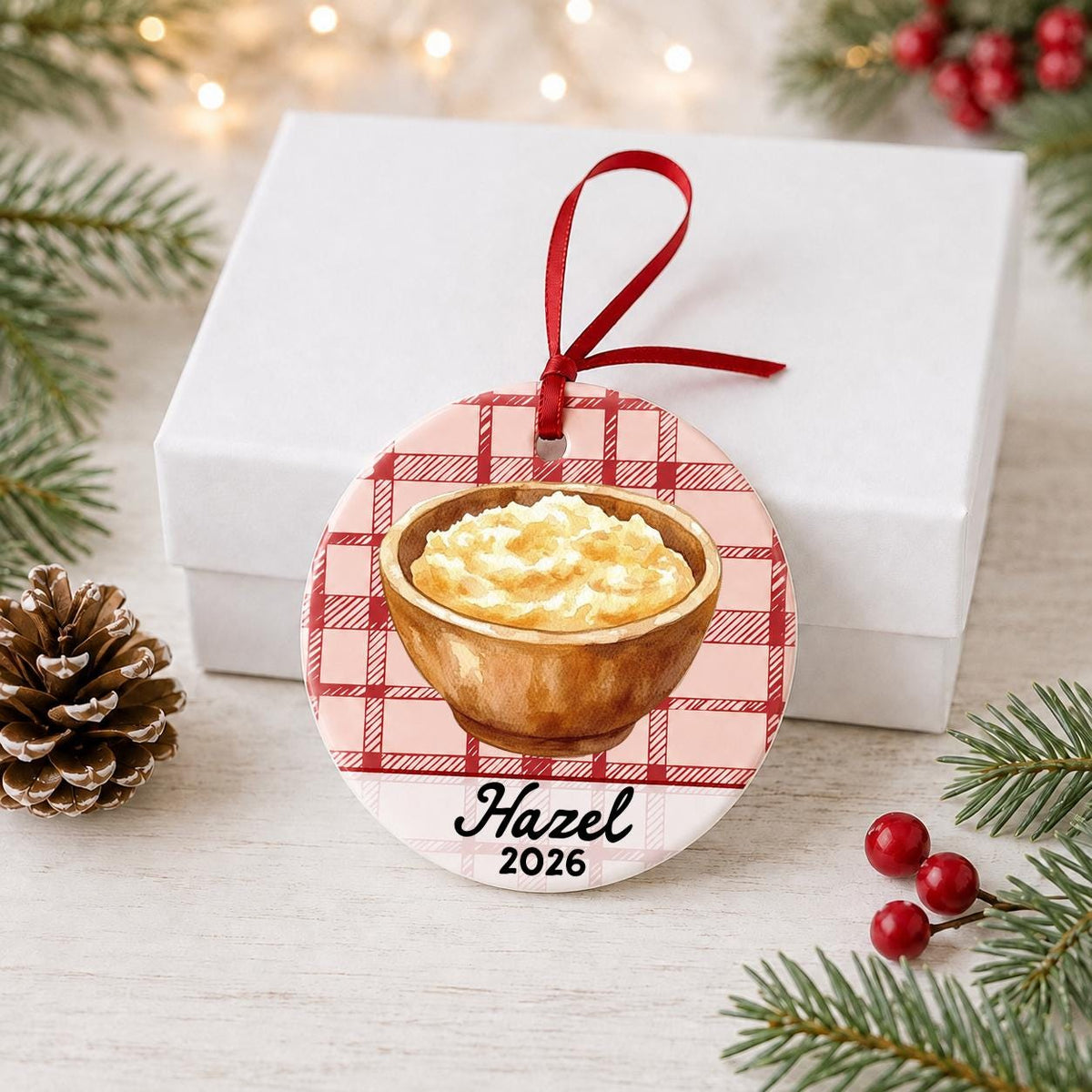 Personalized mashed potatoes ceramic ornament with name and year on red plaid in front of a white gift box with pinecones and red berries