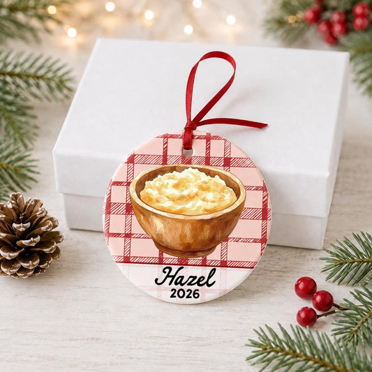 Personalized mashed potatoes ceramic ornament with name and year on red plaid in front of a white gift box with pinecones and red berries