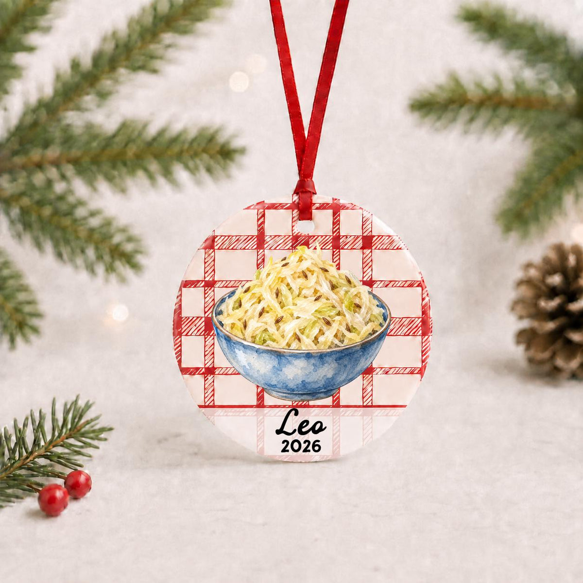 Personalized sauerkraut ceramic ornament with name and year on red plaid with red ribbon on a white surface with pine branches and pinecones
