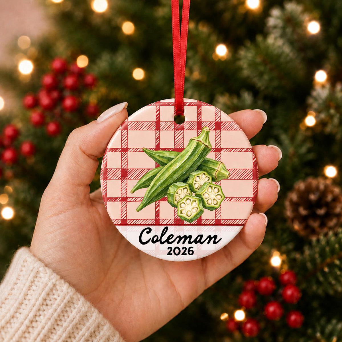Hand holding the personalized okra ceramic ornament with name and year on red plaid in front of a Christmas tree