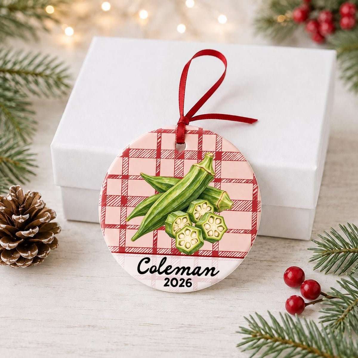 Personalized okra ceramic ornament with name and year on red plaid in front of a white gift box with pinecones and red berries