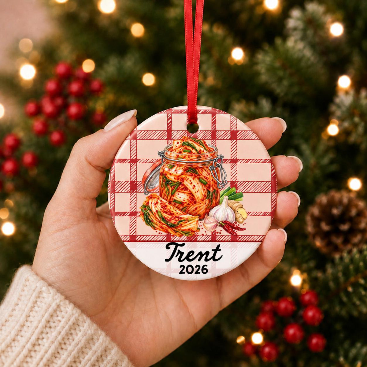 Hand holding the personalized kimchi ceramic ornament with name and year on red plaid in front of a Christmas tree