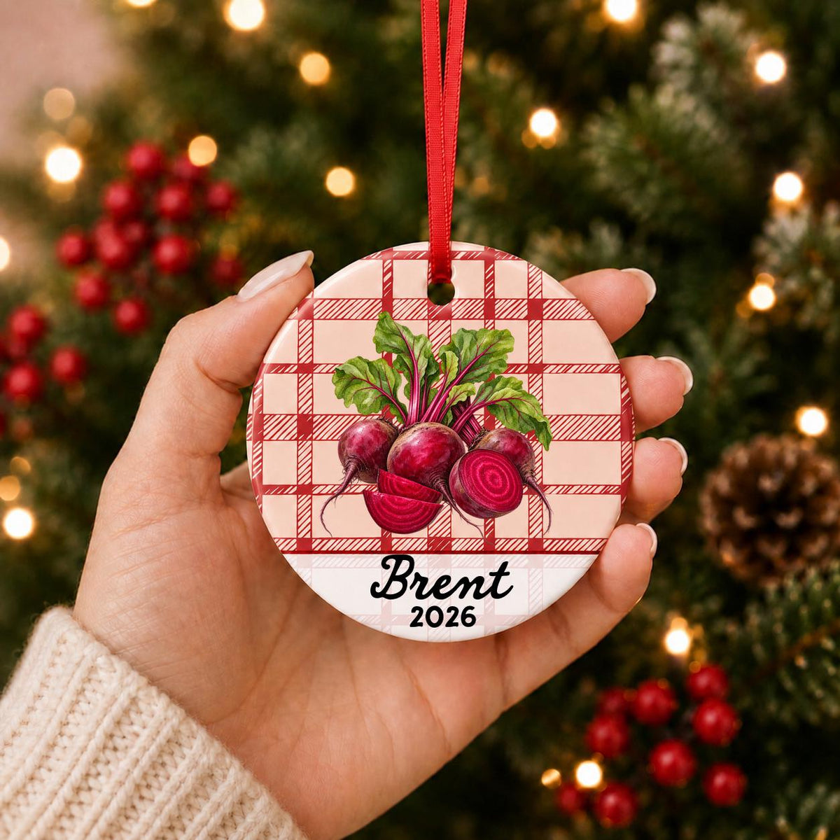 Hand holding the personalized beets ceramic ornament with name and year on red plaid in front of a Christmas tree