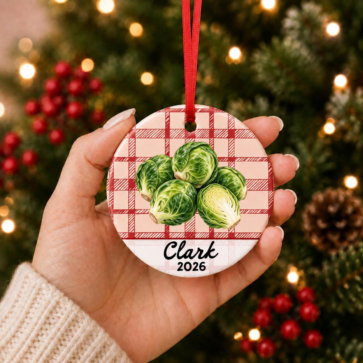 Hand holding the personalized brussels sprouts ceramic ornament with name and year on red plaid in front of a Christmas tree