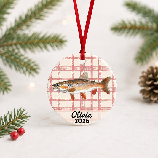 Personalized rainbow trout ceramic ornament with name and year on red plaid with red ribbon on a white surface with pine branches and pinecones