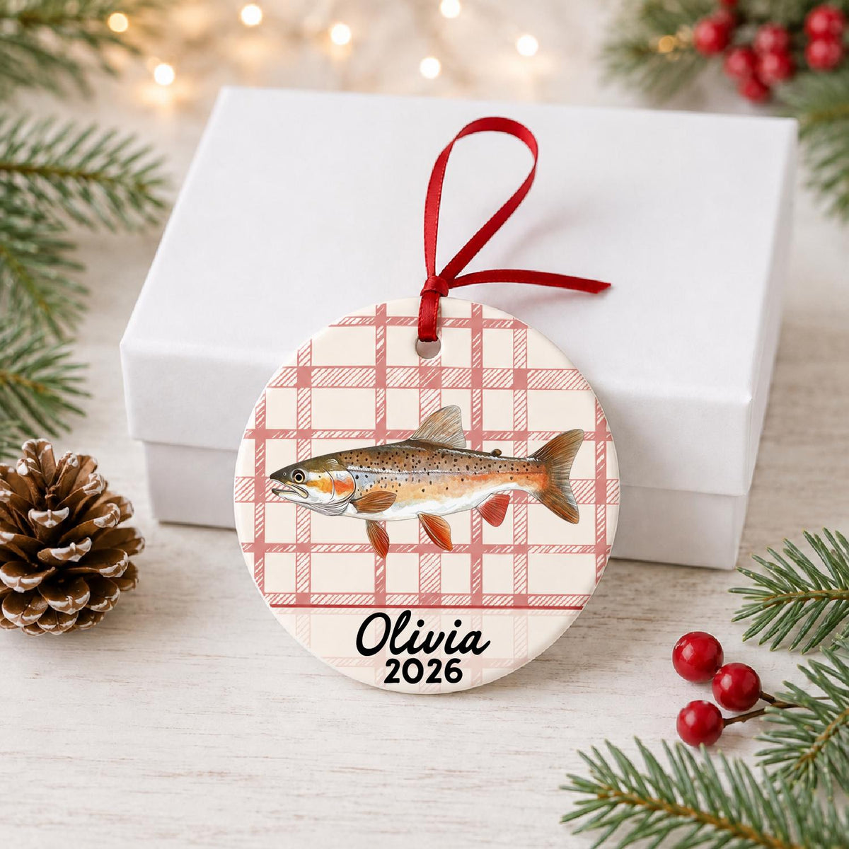Personalized rainbow trout ceramic ornament with name and year on red plaid in front of a white gift box with pinecones and red berries
