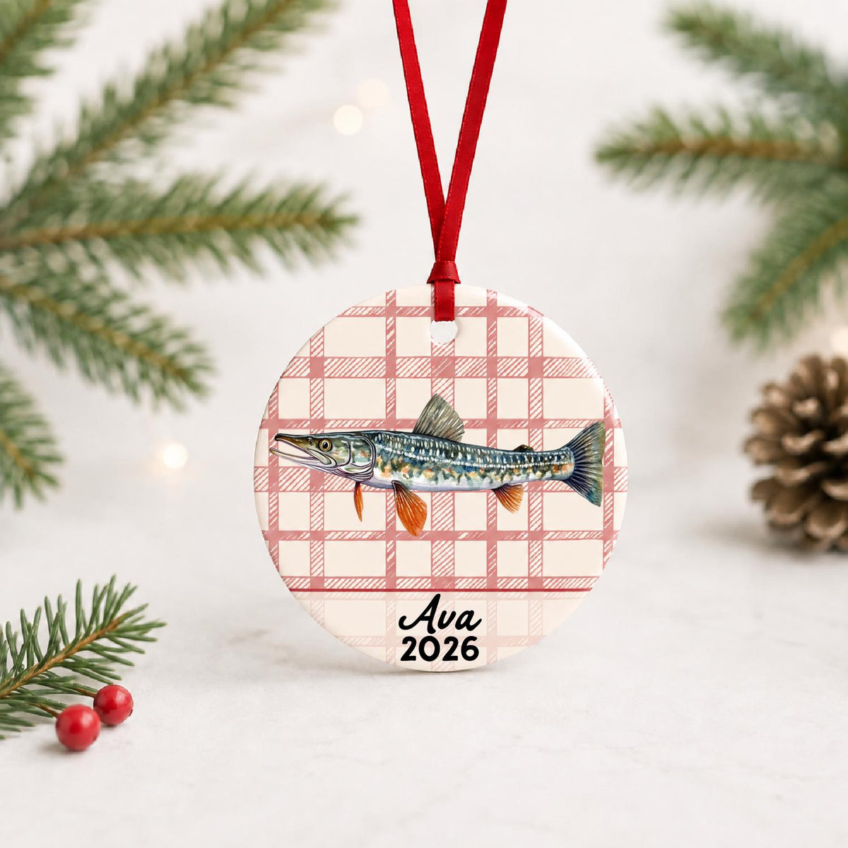 Personalized northern pike ceramic ornament with name and year on red plaid with red ribbon on a white surface with pine branches and pinecones