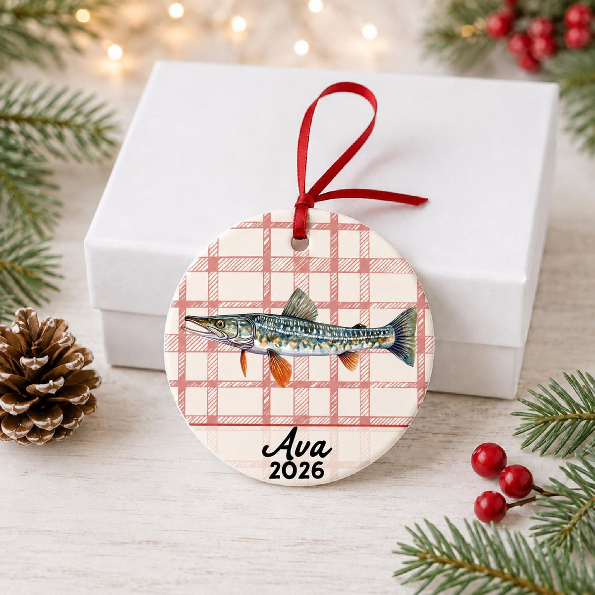 Personalized northern pike ceramic ornament with name and year on red plaid in front of a white gift box with pinecones and red berries