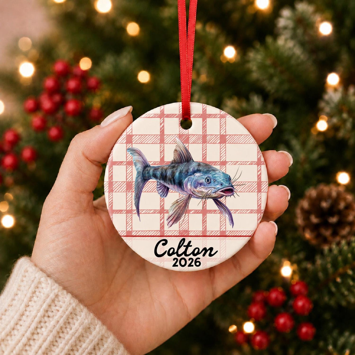 Hand holding the personalized catfish ceramic ornament with name and year on red plaid in front of a Christmas tree