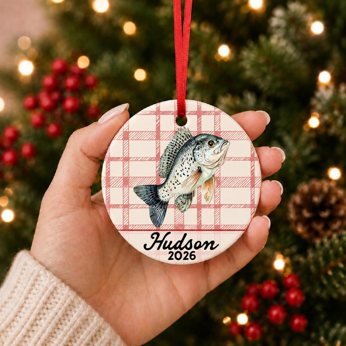 Hand holding the personalized crappie ceramic ornament with name and year on red plaid in front of a Christmas tree