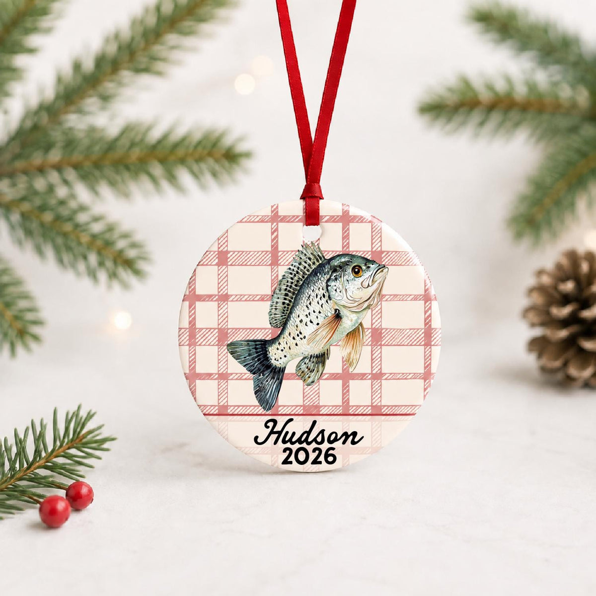 Personalized crappie ceramic ornament with name and year on red plaid with red ribbon on a white surface with pine branches and pinecones