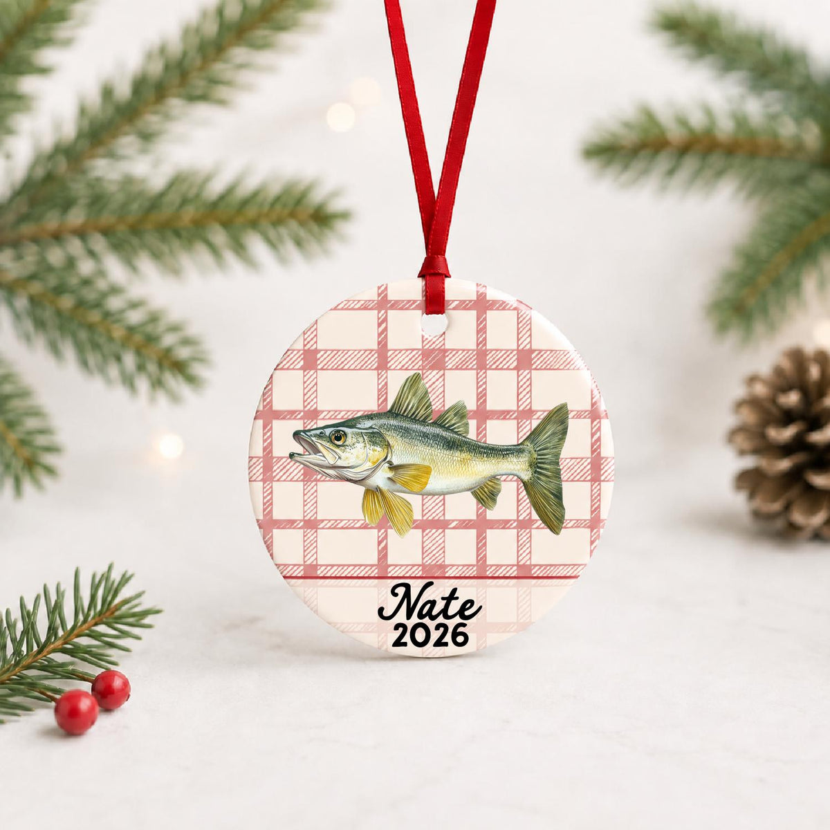 Personalized walleye ceramic ornament with name and year on red plaid with red ribbon on a white surface with pine branches and pinecones