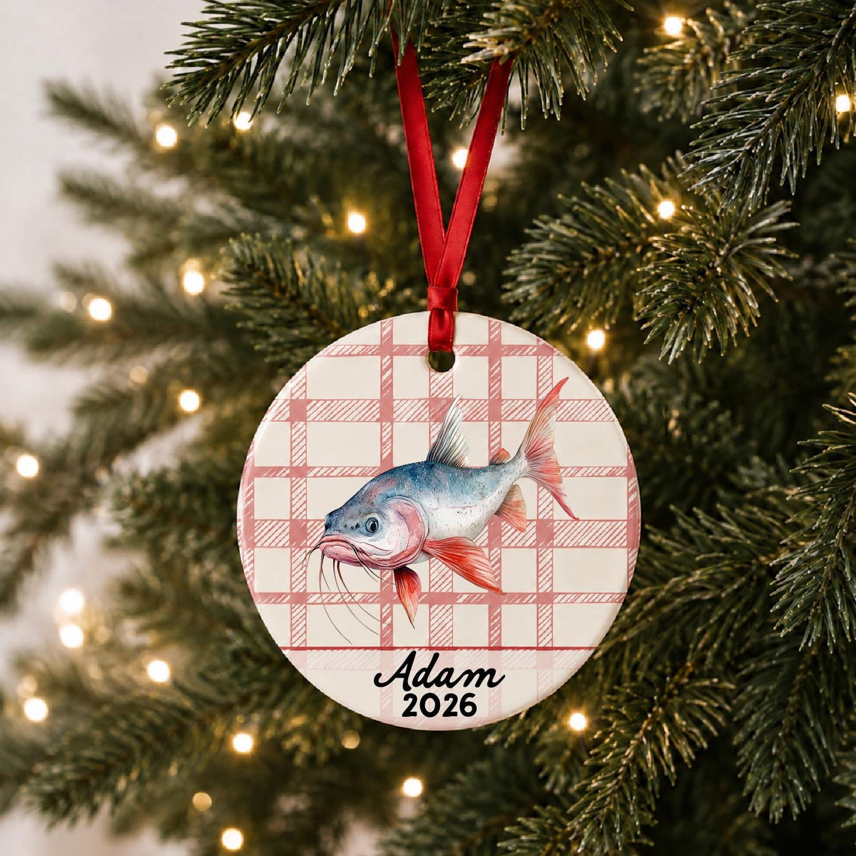 Personalized red-finned catfish ceramic ornament with name and year on red plaid hanging on a lit Christmas tree