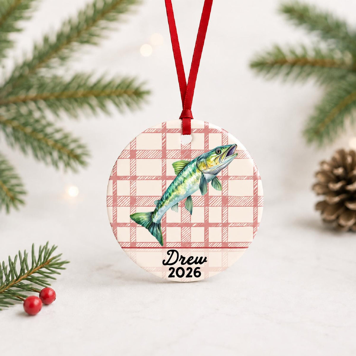 Personalized muskie ceramic ornament with name and year on red plaid with red ribbon on a white surface with pine branches and pinecones