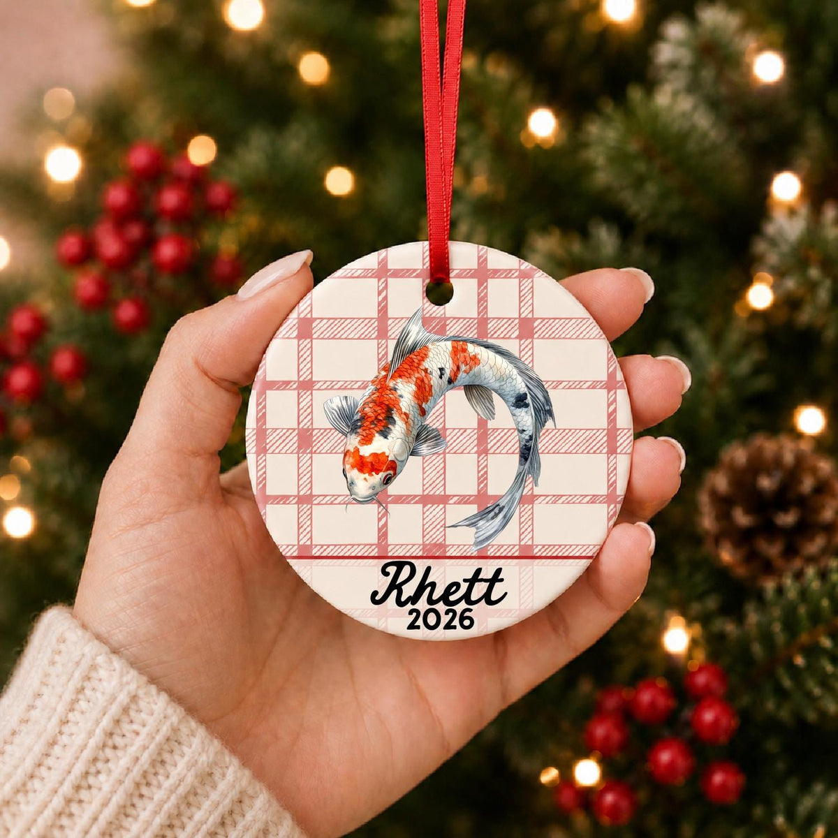 Hand holding the personalized koi fish ceramic ornament with name and year on red plaid in front of a Christmas tree
