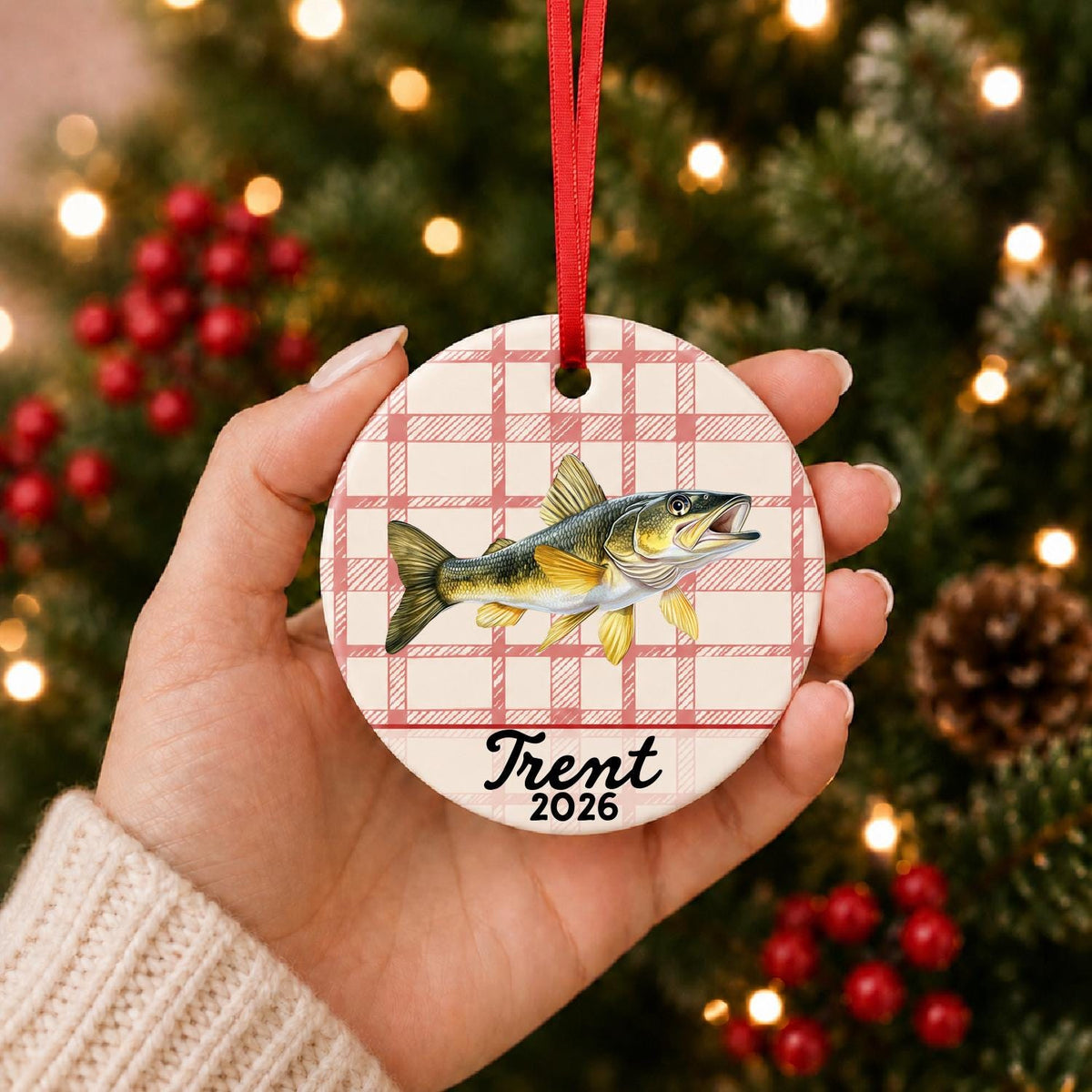Hand holding the personalized golden walleye ceramic ornament with name and year on red plaid in front of a Christmas tree