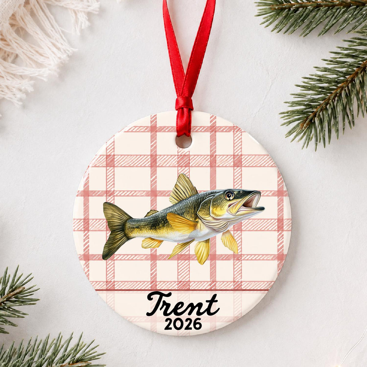 Personalized golden walleye ceramic ornament with name and year on red plaid styled with pine branches on a soft white background