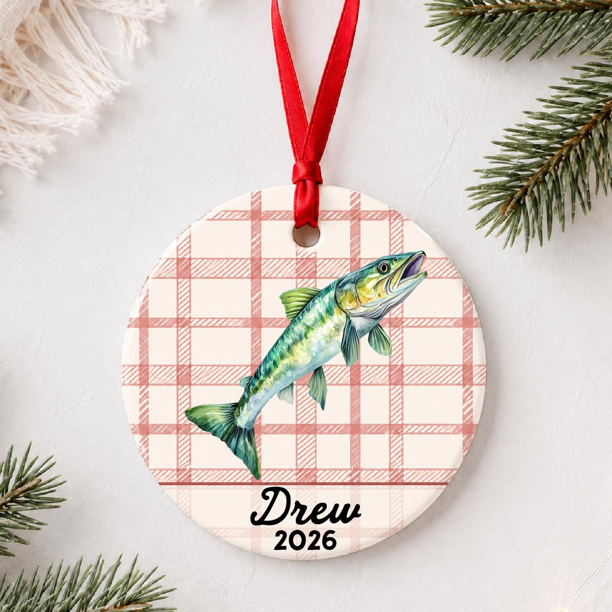 Personalized muskie ceramic ornament with name and year on red plaid styled with pine branches on a soft white background