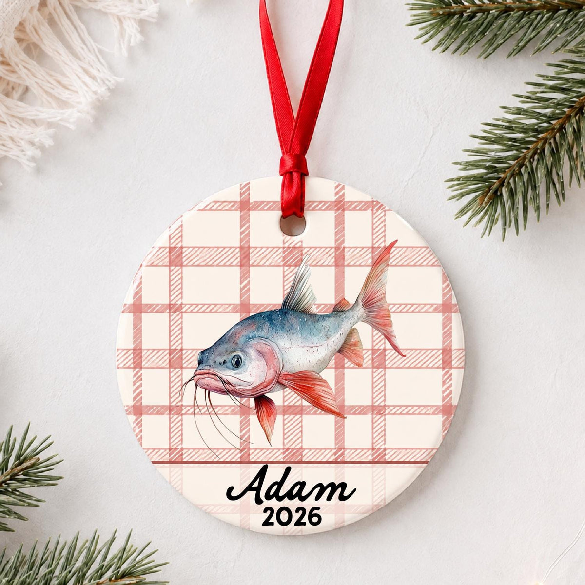 Personalized red-finned catfish ceramic ornament with name and year on red plaid styled with pine branches on a soft white background