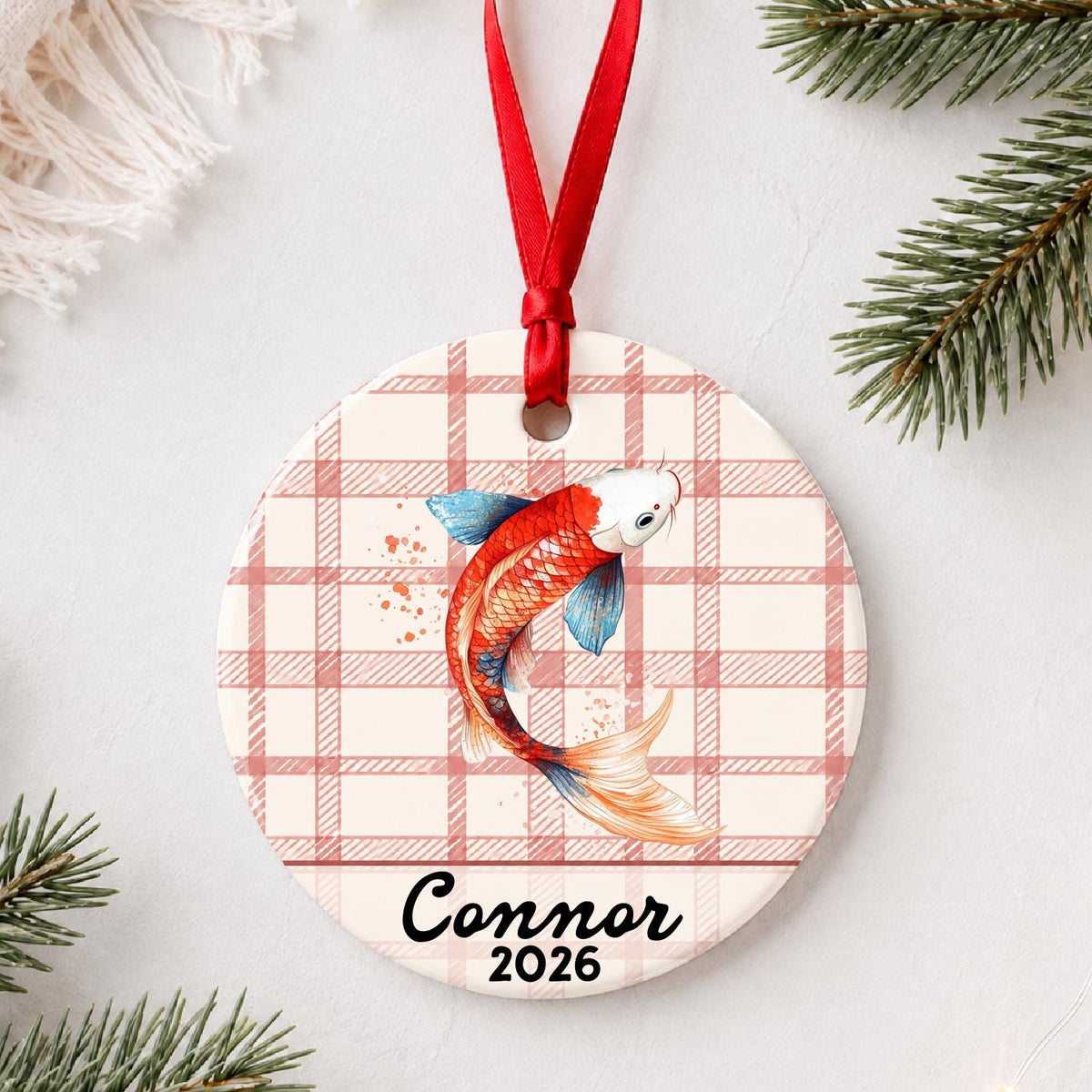 Personalized red and white koi ceramic ornament with name and year on red plaid styled with pine branches on a soft white background