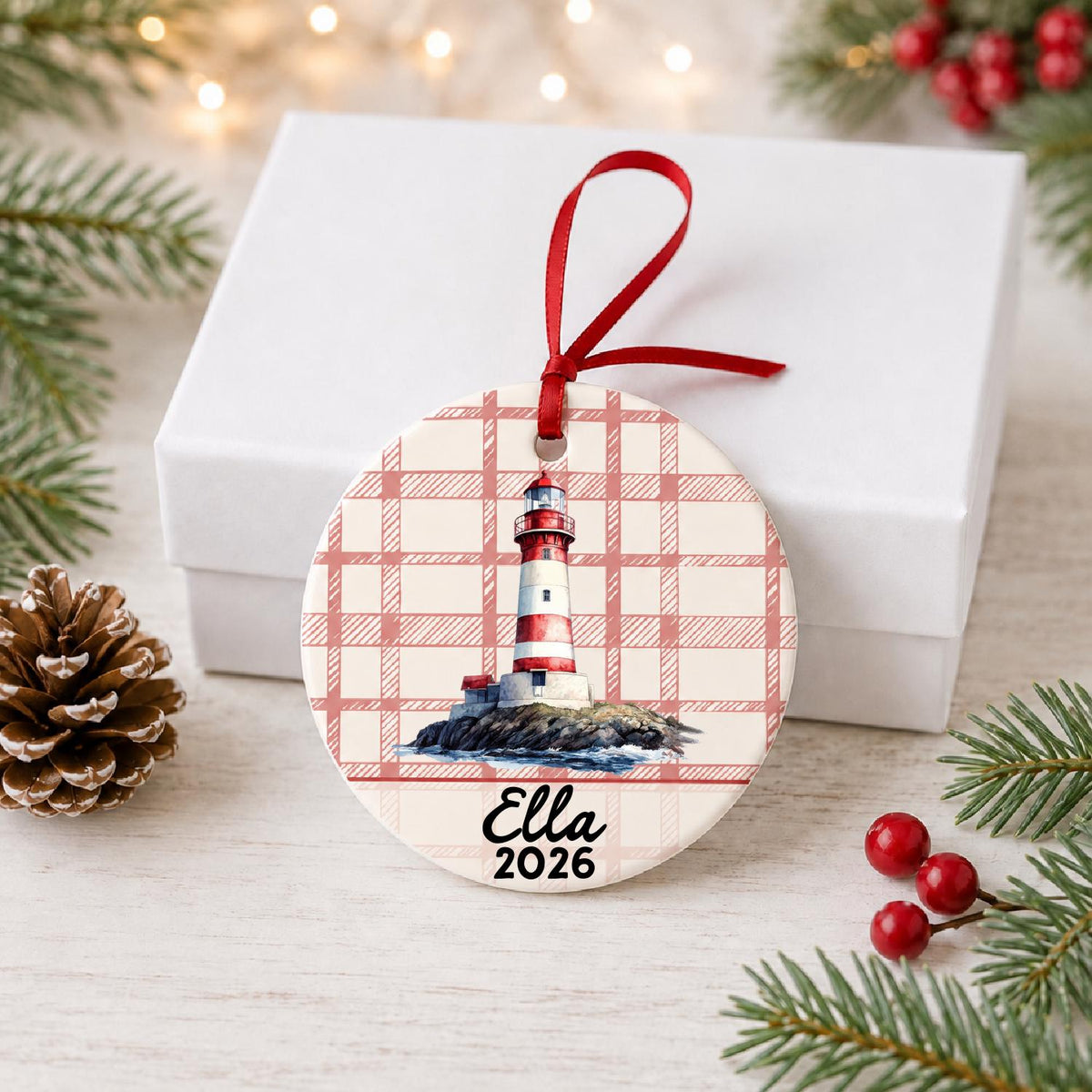 Personalized island lighthouse ceramic ornament with name and year on red plaid in front of a white gift box with pinecones and red berries