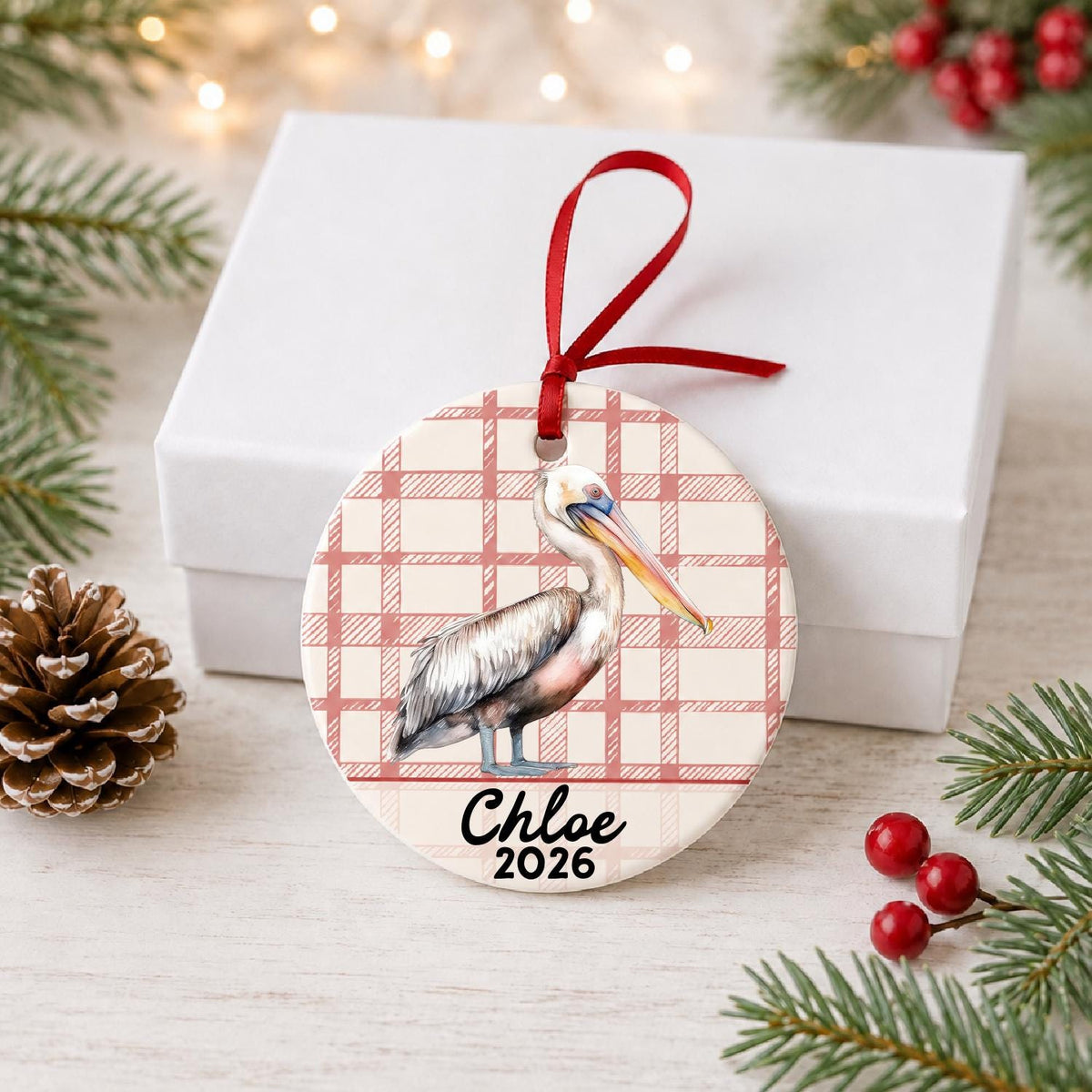 Personalized pelican ceramic ornament with name and year on red plaid in front of a white gift box with pinecones and red berries
