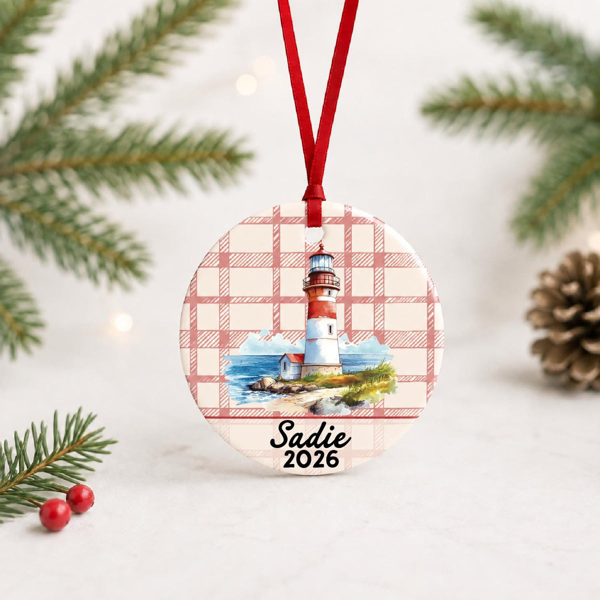 Personalized beach lighthouse ceramic ornament with name and year on red plaid with red ribbon on a white surface with pine branches and pinecones