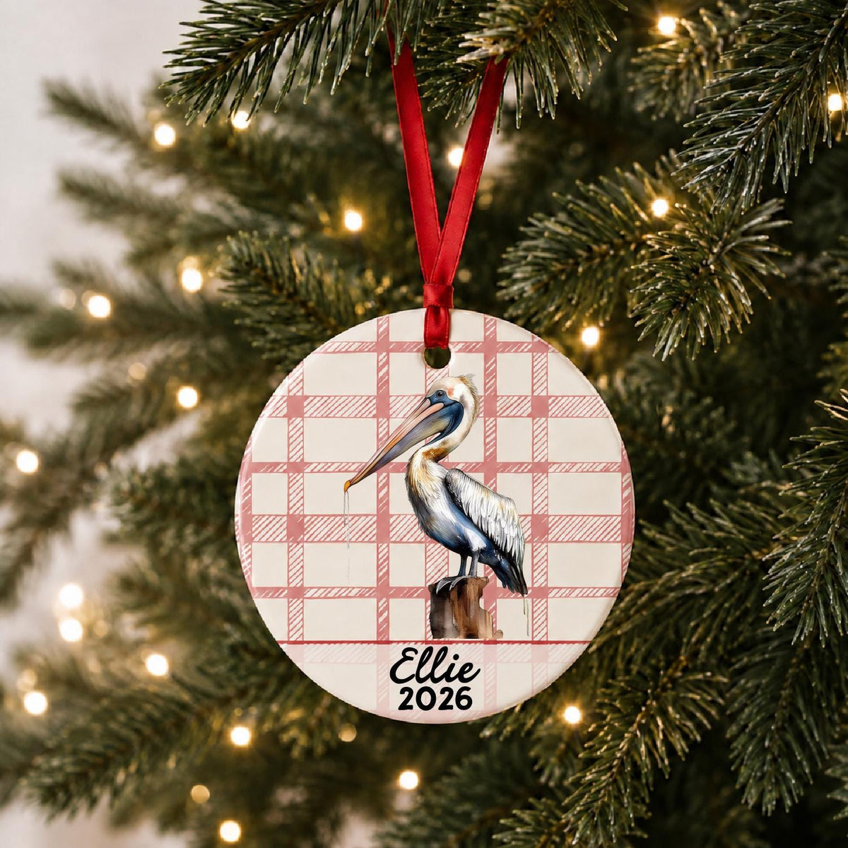 Personalized pelican on piling ceramic ornament with name and year on red plaid hanging on a lit Christmas tree