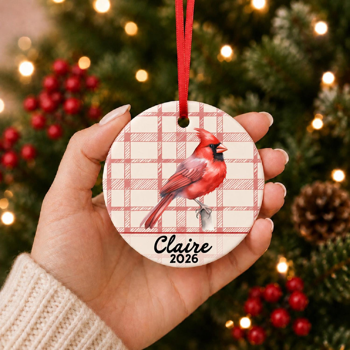 Hand holding the personalized northern cardinal ceramic ornament with name and year on red plaid in front of a Christmas tree
