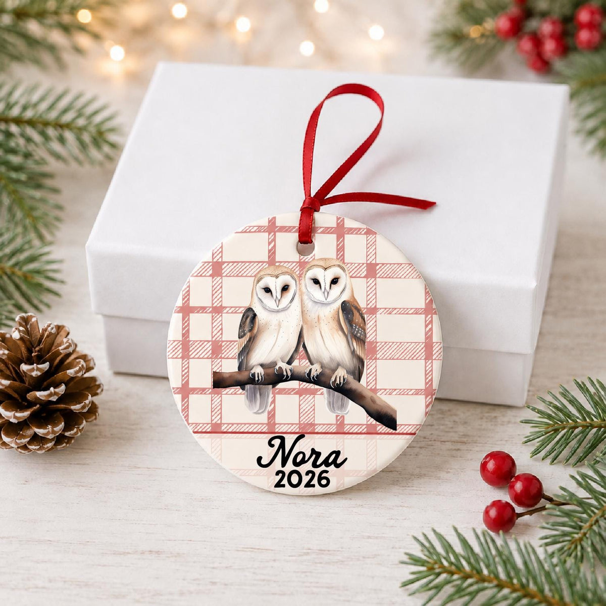 Personalized barn owl pair ceramic ornament with name and year on red plaid in front of a white gift box with pinecones and red berries