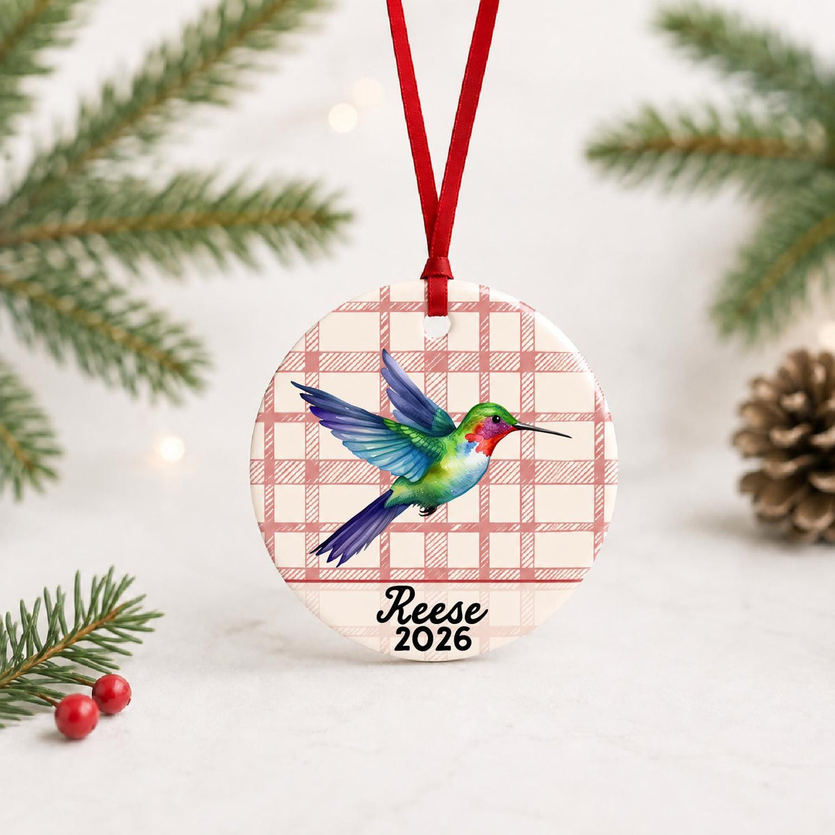 Personalized hummingbird ceramic ornament with name and year on red plaid with red ribbon on a white surface with pine branches and pinecones