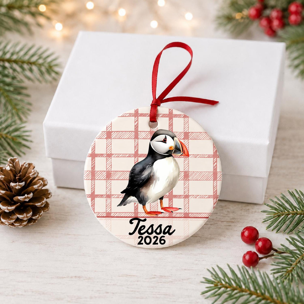 Personalized puffin ceramic ornament with name and year on red plaid in front of a white gift box with pinecones and red berries