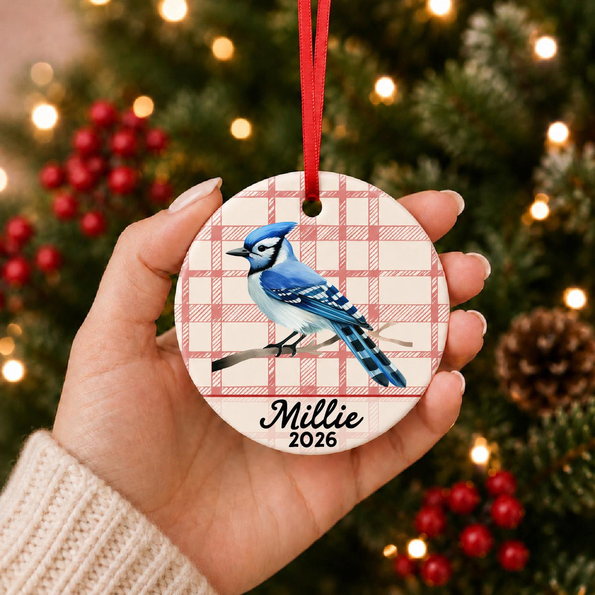 Hand holding the personalized blue jay ceramic ornament with name and year on red plaid in front of a Christmas tree