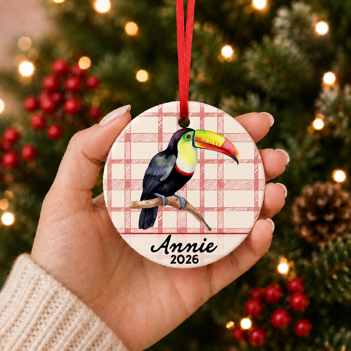 Hand holding the personalized toucan ceramic ornament with name and year on red plaid in front of a Christmas tree