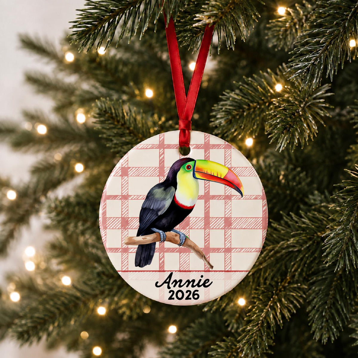 Personalized toucan ceramic ornament with name and year on red plaid hanging on a lit Christmas tree