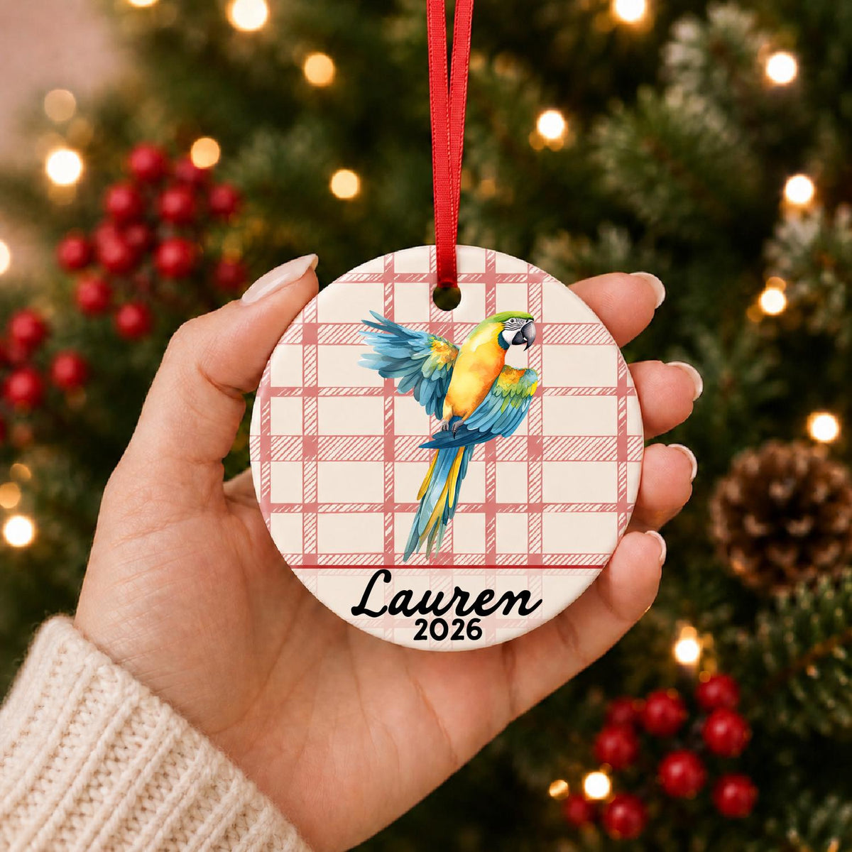 Hand holding the personalized blue and gold macaw ceramic ornament with name and year on red plaid in front of a Christmas tree