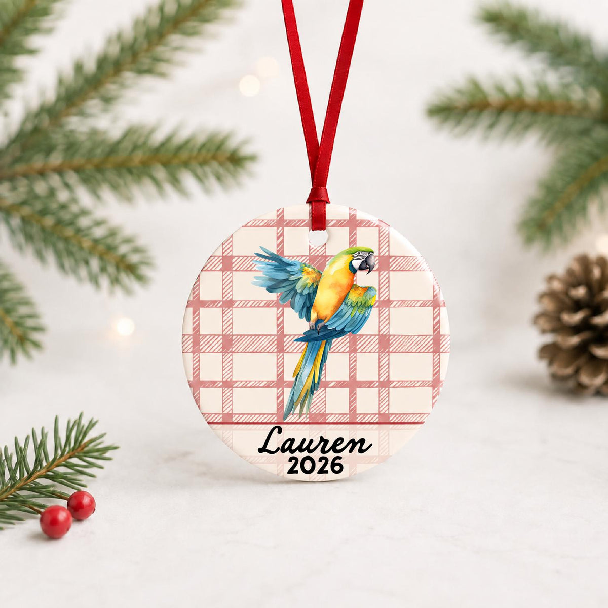Personalized blue and gold macaw ceramic ornament with name and year on red plaid with red ribbon on a white surface with pine branches and pinecones