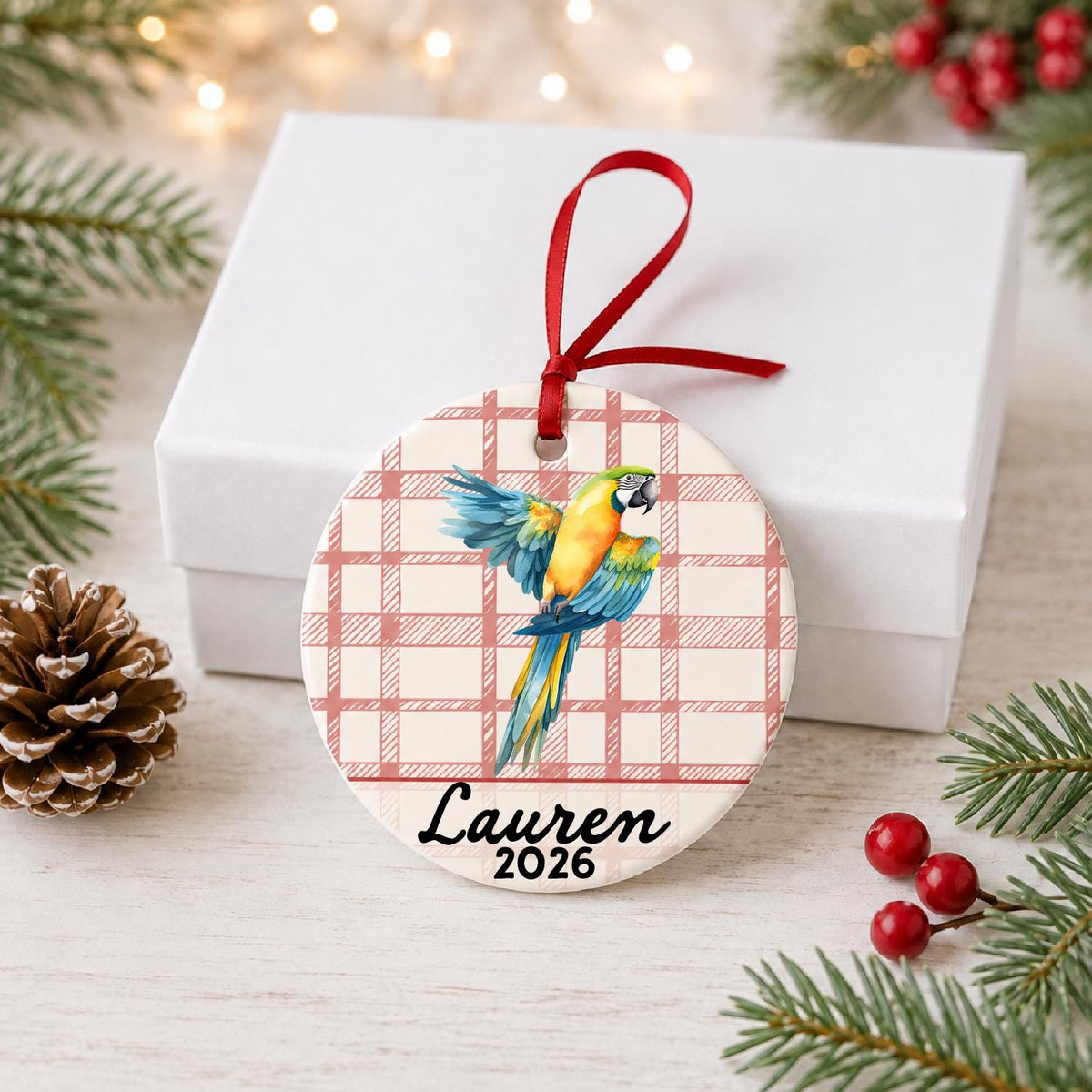 Personalized blue and gold macaw ceramic ornament with name and year on red plaid in front of a white gift box with pinecones and red berries