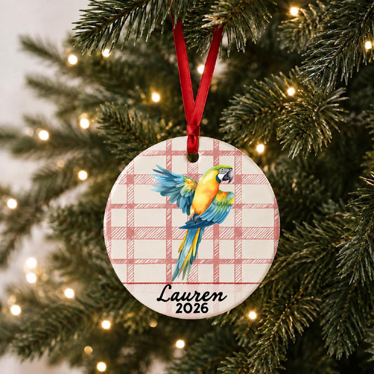 Personalized blue and gold macaw ceramic ornament with name and year on red plaid hanging on a lit Christmas tree