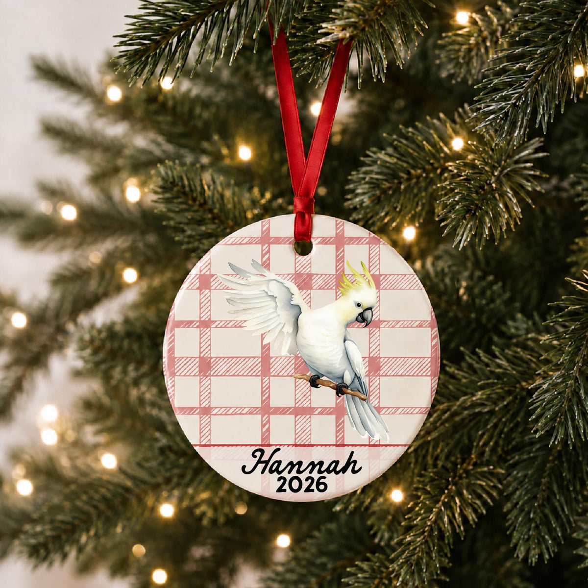 Personalized cockatoo ceramic ornament with name and year on red plaid hanging on a lit Christmas tree