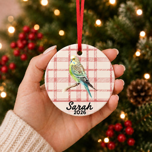 Hand holding the personalized budgerigar ceramic ornament with name and year on red plaid in front of a Christmas tree