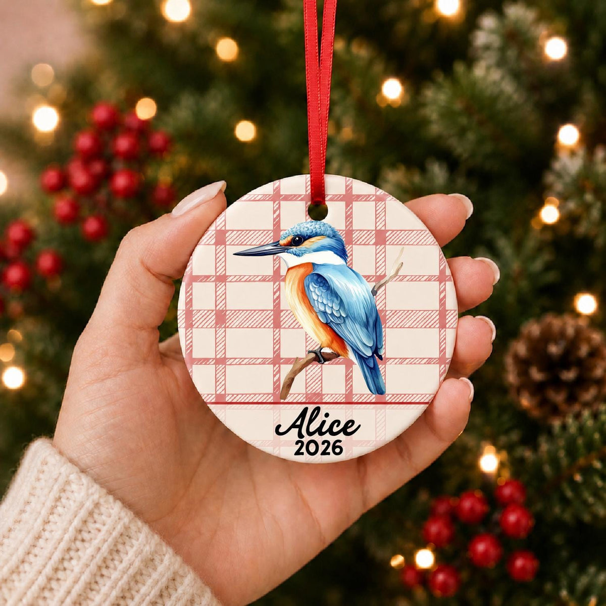 Hand holding the personalized kingfisher ceramic ornament with name and year on red plaid in front of a Christmas tree