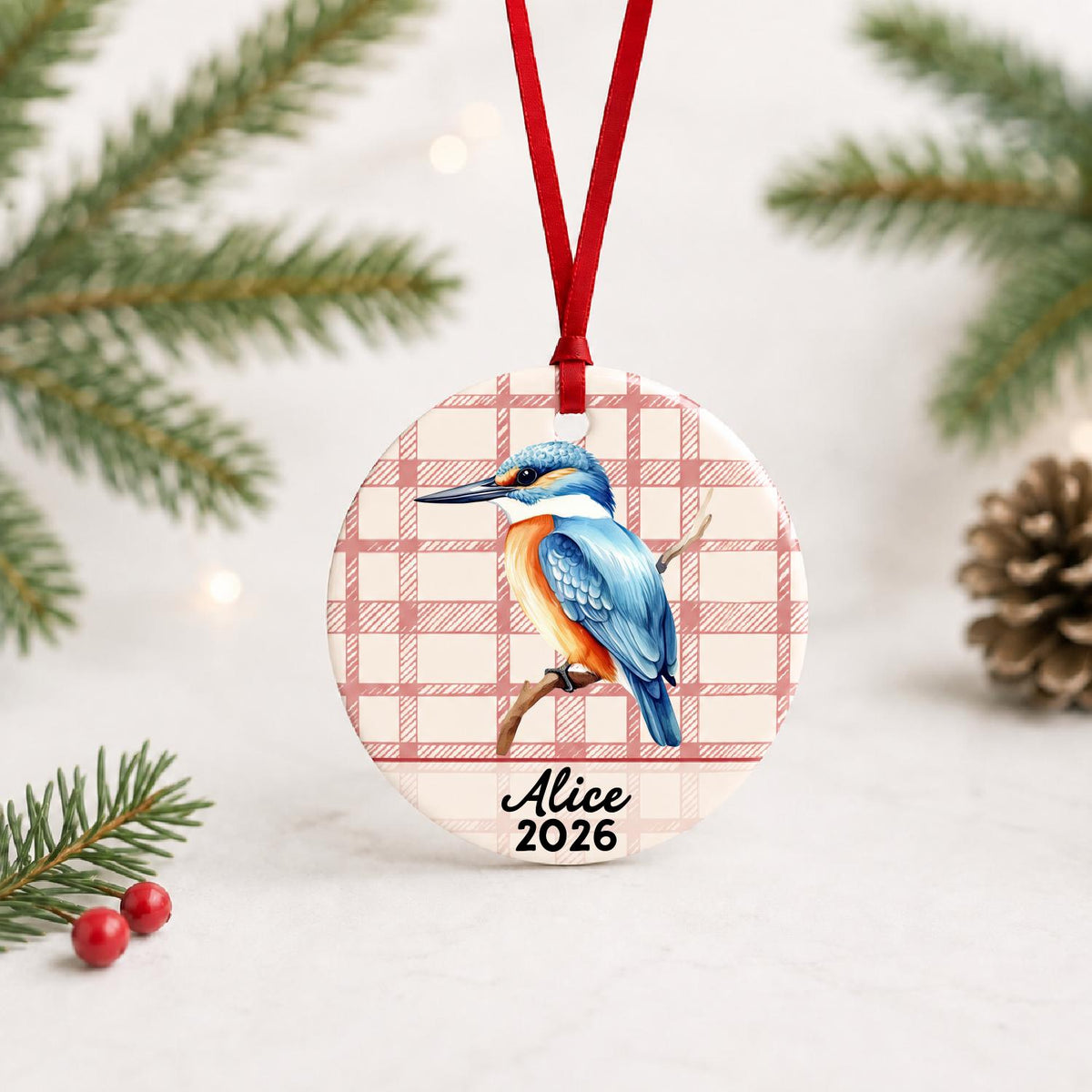 Personalized kingfisher ceramic ornament with name and year on red plaid with red ribbon on a white surface with pine branches and pinecones