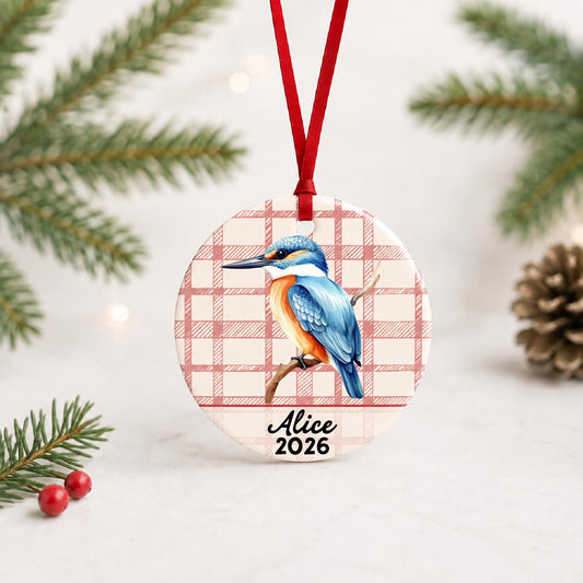Personalized kingfisher ceramic ornament with name and year on red plaid with red ribbon on a white surface with pine branches and pinecones