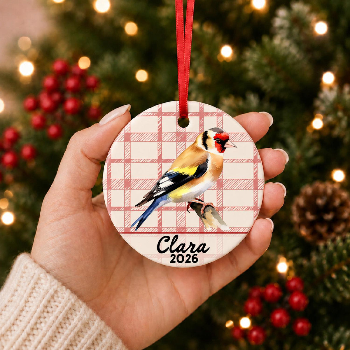 Hand holding the personalized European goldfinch ceramic ornament with name and year on red plaid in front of a Christmas tree
