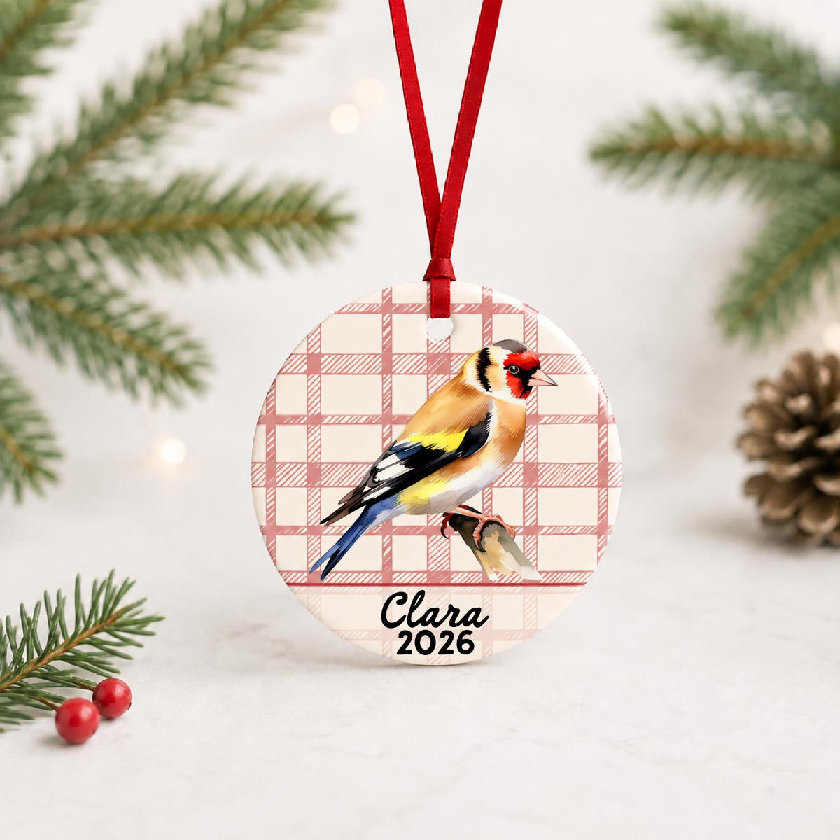 Personalized European goldfinch ceramic ornament with name and year on red plaid with red ribbon on a white surface with pine branches and pinecones