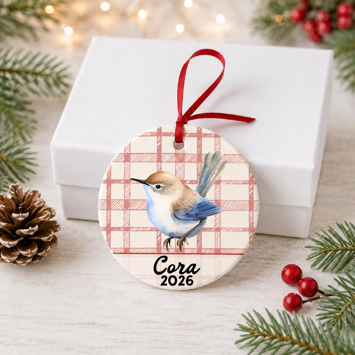 Personalized wren ceramic ornament with name and year on red plaid in front of a white gift box with pinecones and red berries