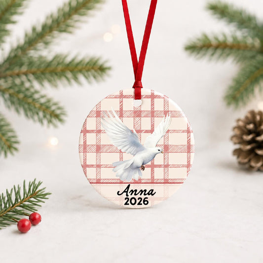 Personalized white dove ceramic ornament with name and year on red plaid with red ribbon on a white surface with pine branches and pinecones