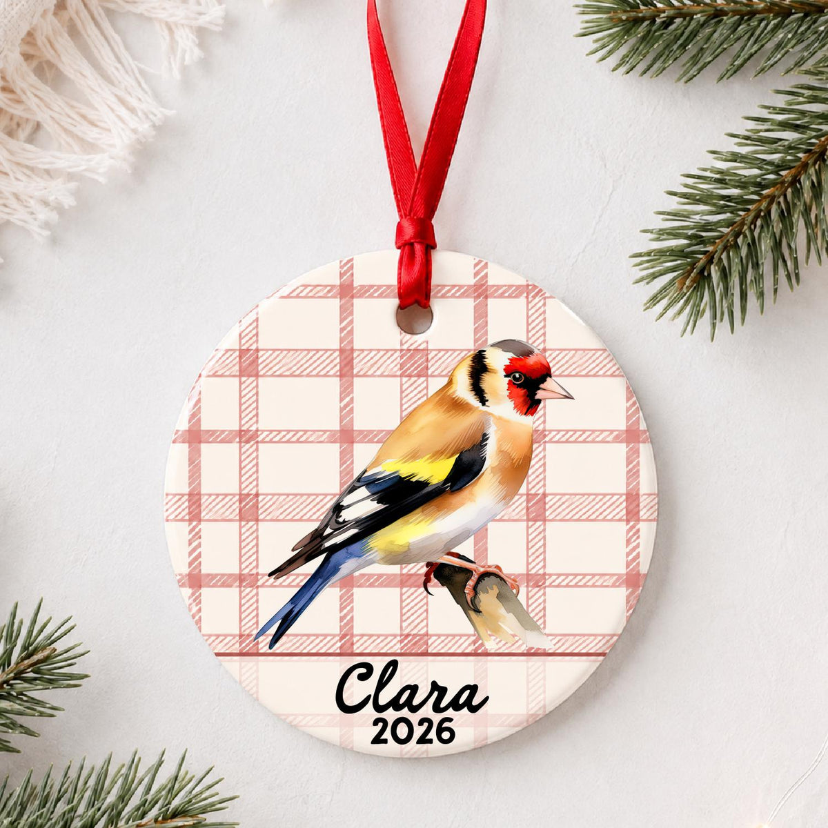 Personalized European goldfinch ceramic ornament with name and year on red plaid styled with pine branches on a soft white background